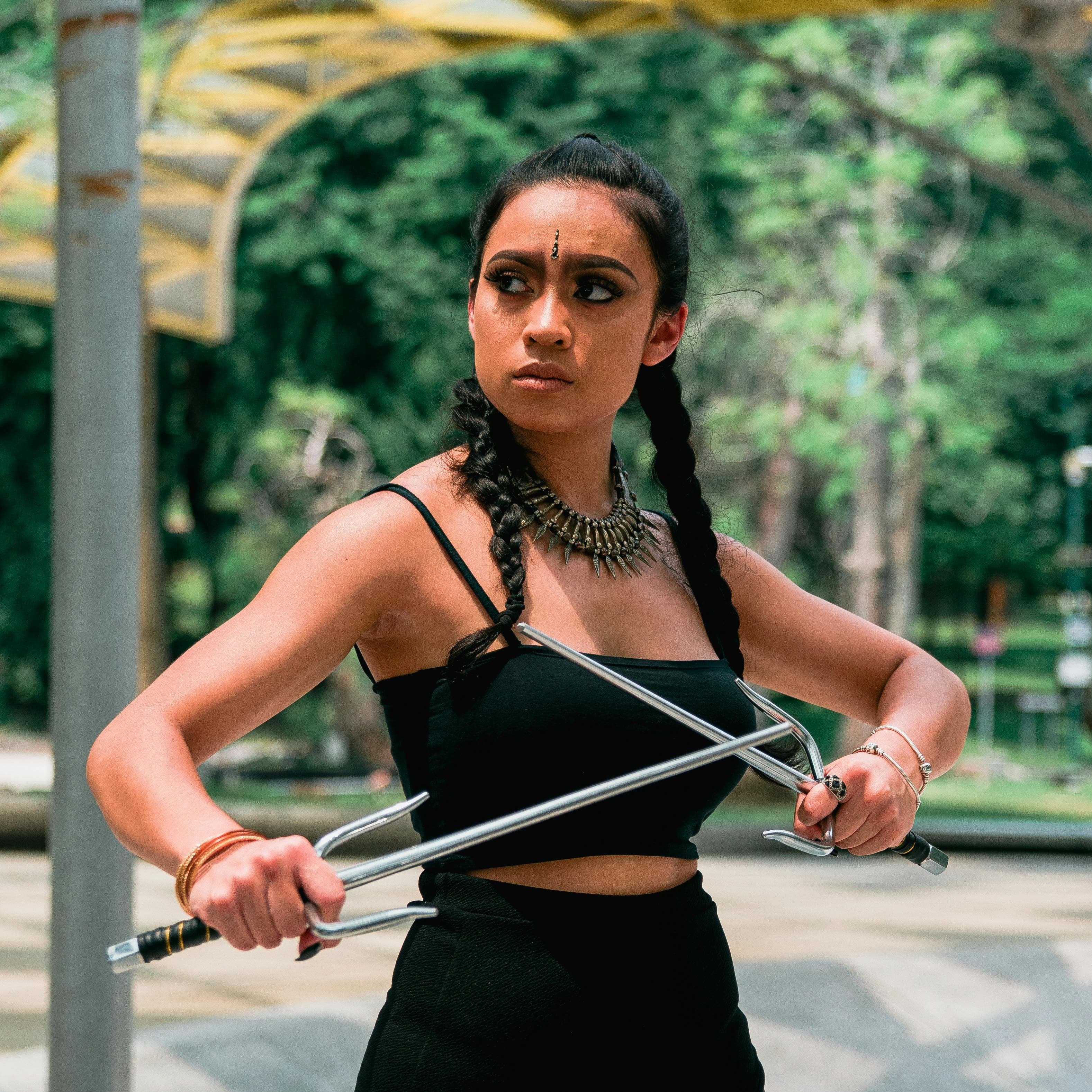 A woman confidently posing with martial arts sai, showcasing beauty and strength, set against an outdoor background.