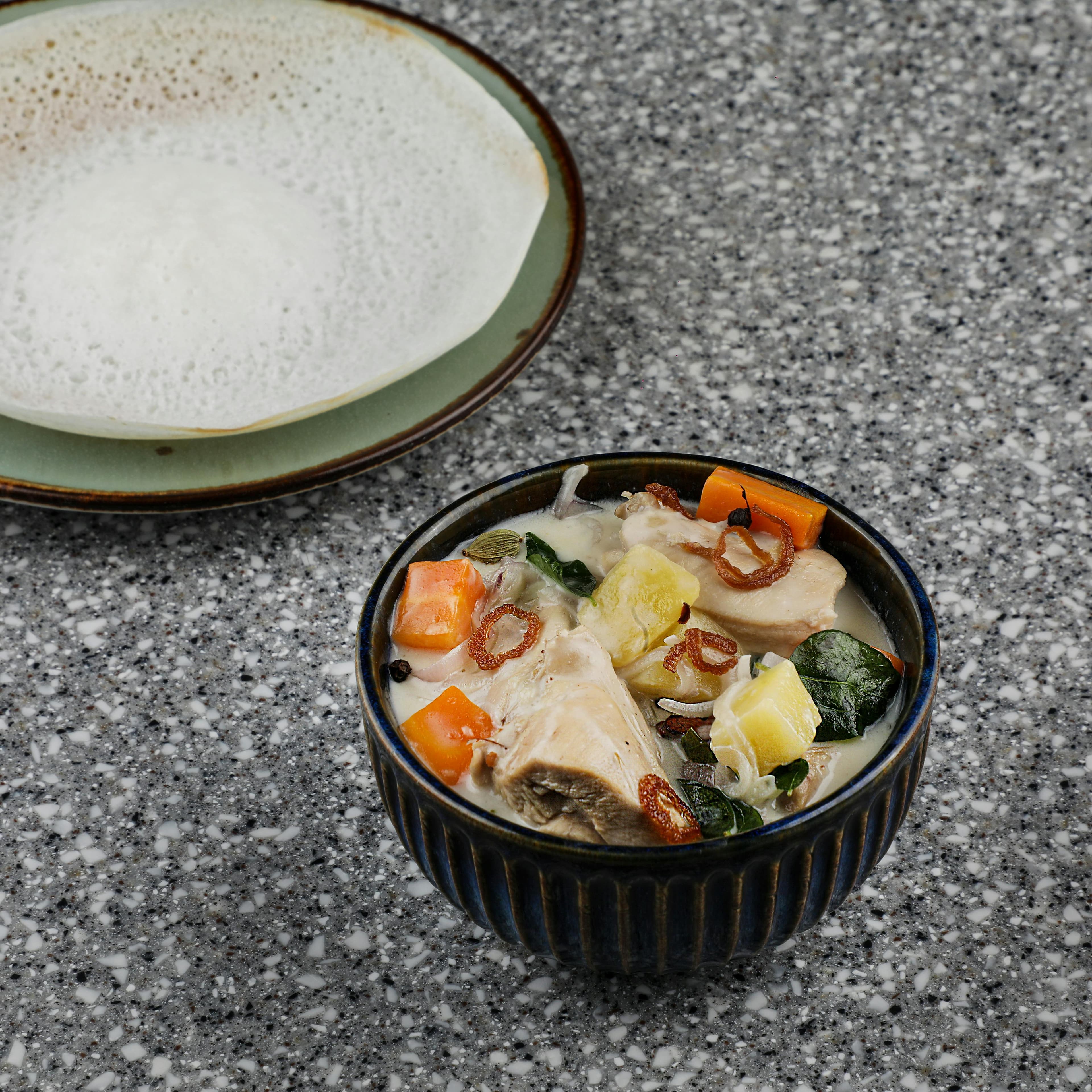 Uma tigela de ensopado de legumes ao estilo de Kerala servido com appam em uma mesa de granito, exibindo a culinária indiana.