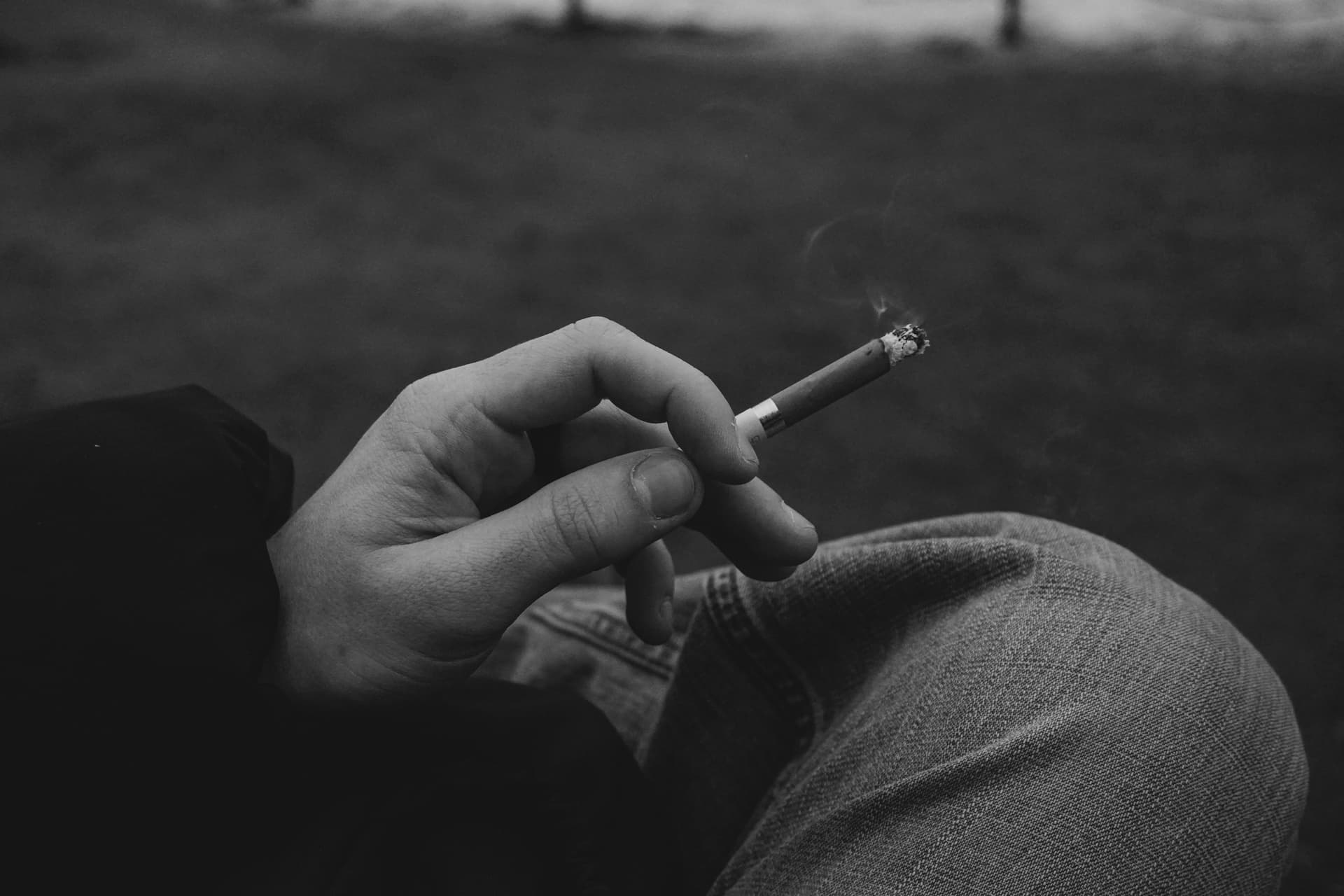 Monochrome image of a hand holding a cigarette outdoors. Smoke rises in a calm, contemplative mood.