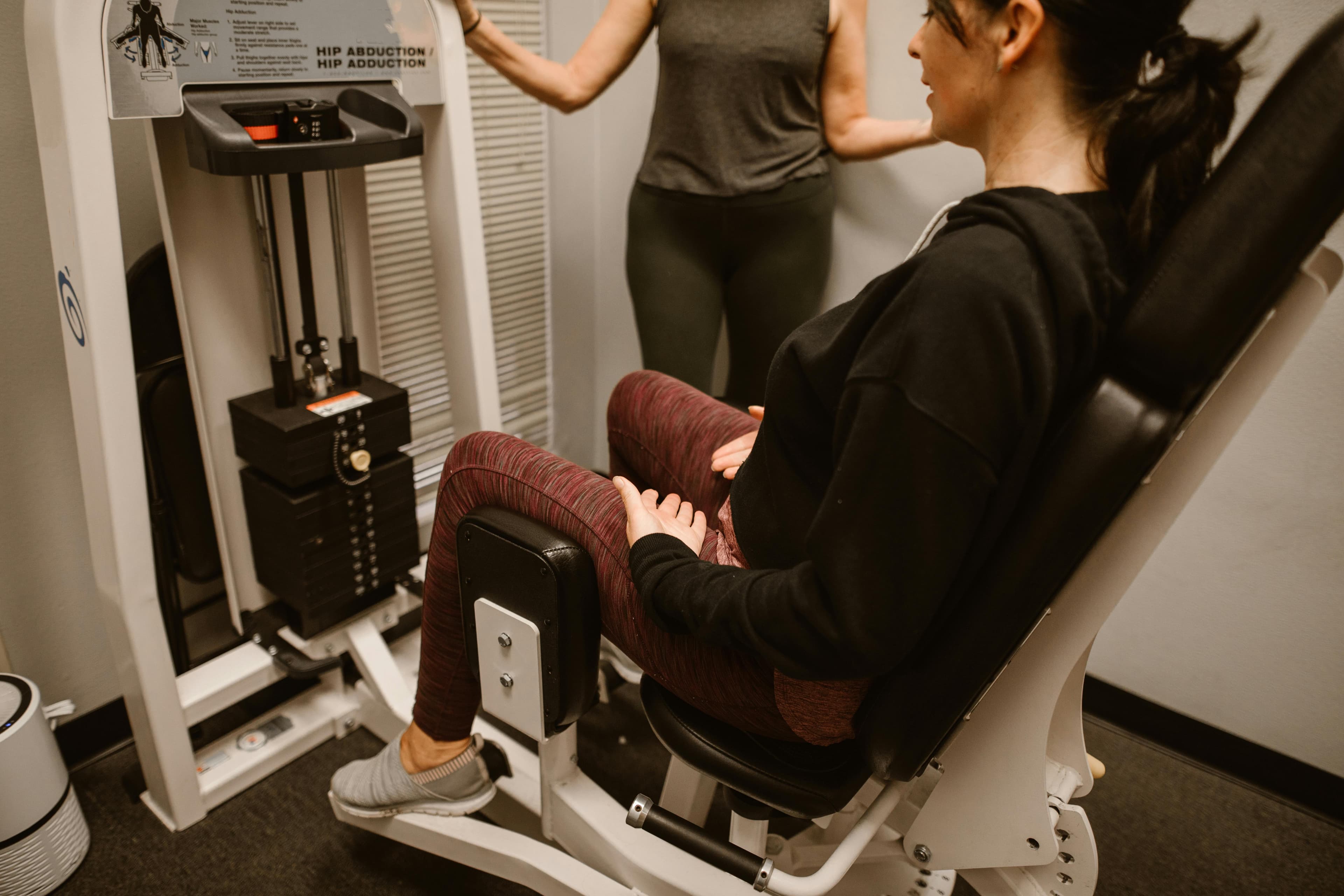 A woman performs exercises on a hip abductor machine with guidance from a trainer.