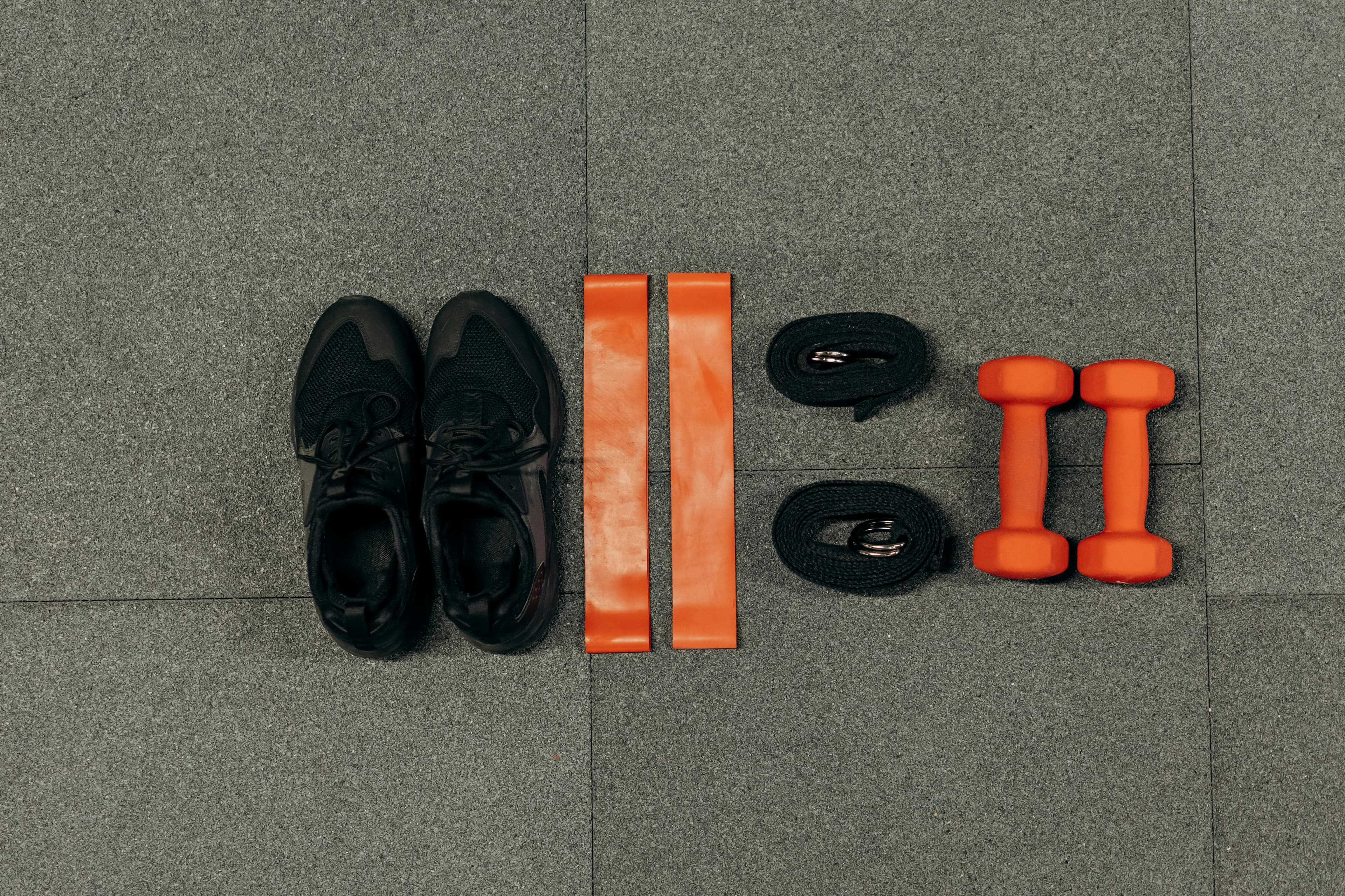 Flat lay of gym essentials including dumbbells, sneakers, and resistance bands on a grey floor.
