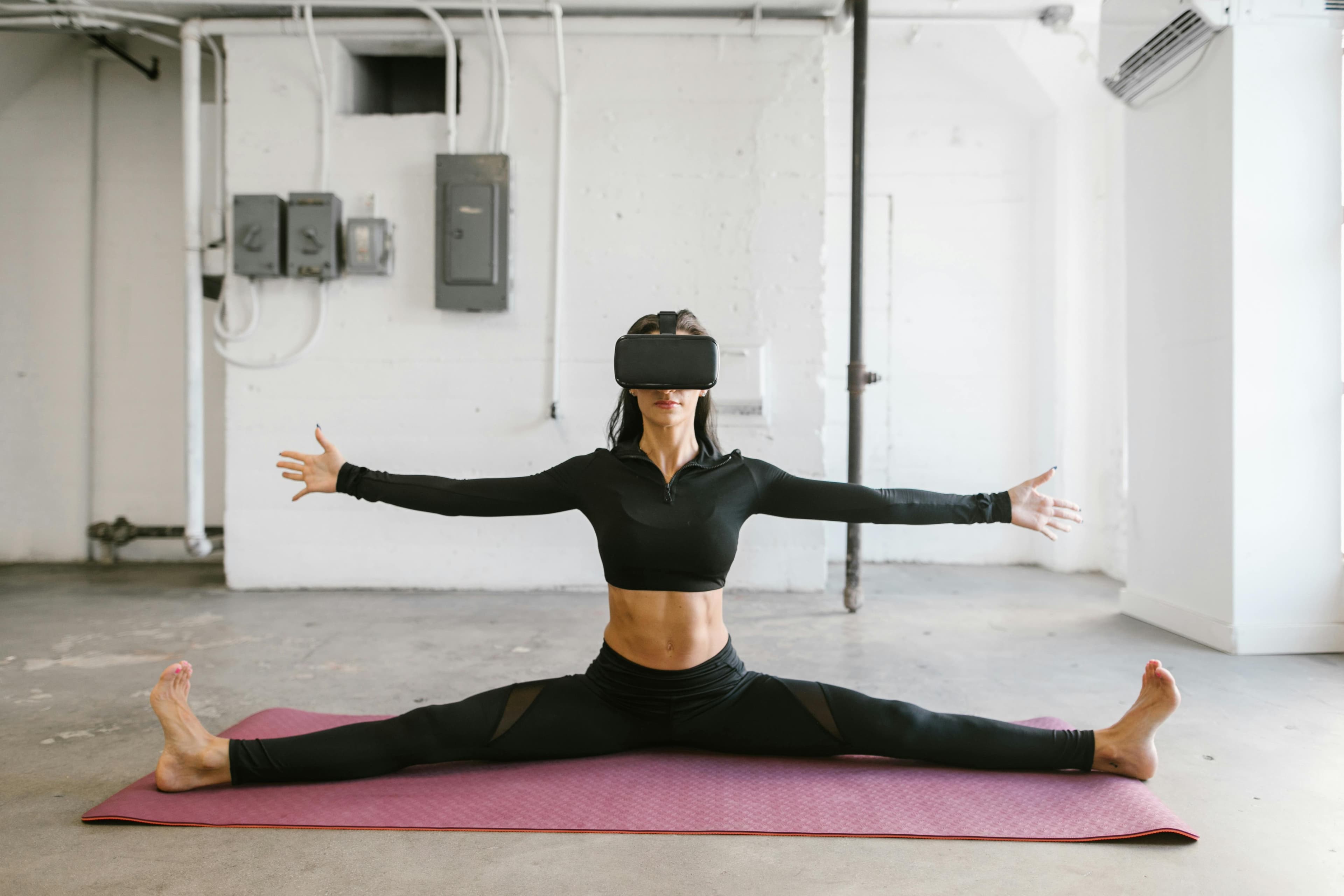 A woman stretches on a yoga mat while wearing a virtual reality headset, indoors.